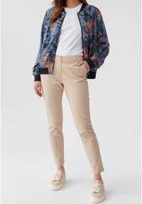 Květinová bomber bunda s modrým podkladem, černé manžety; bílá košile; béžové slim kalhoty; béžové tenisky s texturovanými akcenty.