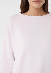 Jasnoróżowy, oversize'owy sweatshirt z szerokim okrągłym dekoltem i gładką fakturą. Posiada opadające ramiona oraz subtelne detale szwów.