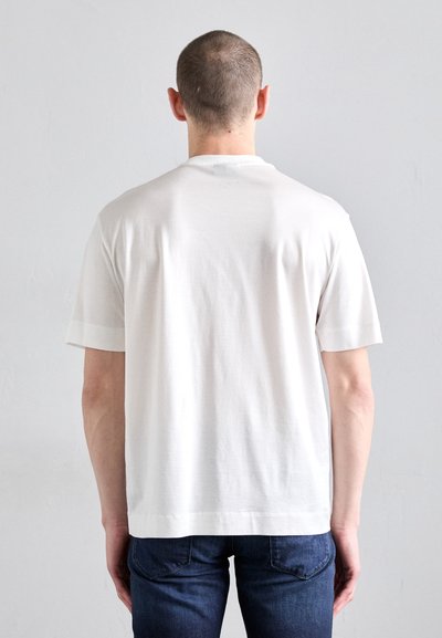 T-shirt en coton blanc à manches courtes, col rond et coupe décontractée, porté avec un jeans bleu foncé. Texture lisse, design minimal.
