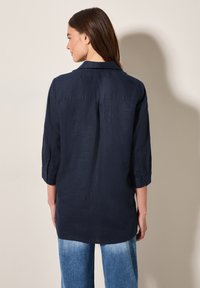 Camicia di lino blu navy con colletto e maniche arrotolate, caratterizzata da un orlo dritto e una texture sottile, indossata con pantaloni in denim chiaro.