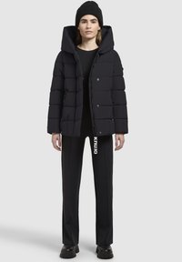Schwarze Daunenjacke mit einer großen Kapuze, die mit gesteppten Abschnitten, vorderen Knöpfen und Reißverschlusstaschen ausgestattet ist, kombiniert mit schwarzen Hosen und einer Mütze.