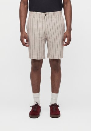 Mann trägt beige Shorts mit dünnen vertikalen Streifen, weiße Socken und dunkelrote Turnschuhe, steht vor einem einfarbigen weißen Hintergrund.