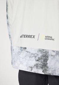 Light grey jacket with a textured pattern at the hem, featuring black "Terrex" and "National Geographic" logos. Smooth fabric finish.
