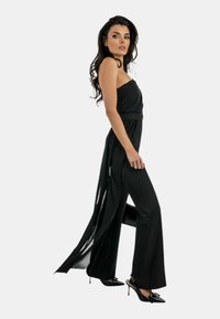 Tuta nera con design senza spalline, vita arricciata e tessuto fluido. Pantaloni svasati e dettagli con spacco accentuano il movimento. Abbinato a tacchi neri.