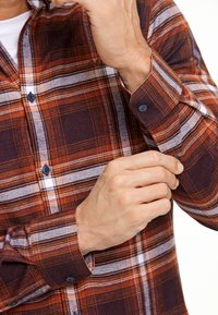 Camicia di flanella a quadri arancione, marrone e bianca. Caratterizzata da una chiusura con bottoni, due bottoni sui polsini e una consistenza morbida.
