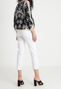 Kvinde iført sort blomstret lace-top, hvidde cropped jeans og sorte høje hæle, stående mod en ensfarvet baggrund.