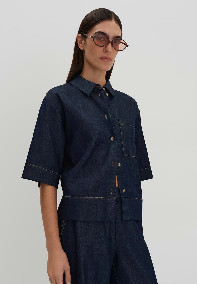 Dark denim short-sleeve button-up shirt with a chest pocket, contrasting yellow stitching, and a relaxed fit. Model wears tinted sunglasses.