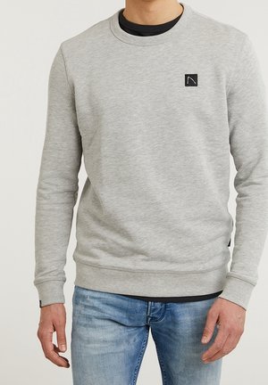 Man met een lichtgrijze sweatshirthals met zwart logopatch op de borst en blauwe jeans, staand tegen een effen achtergrond.