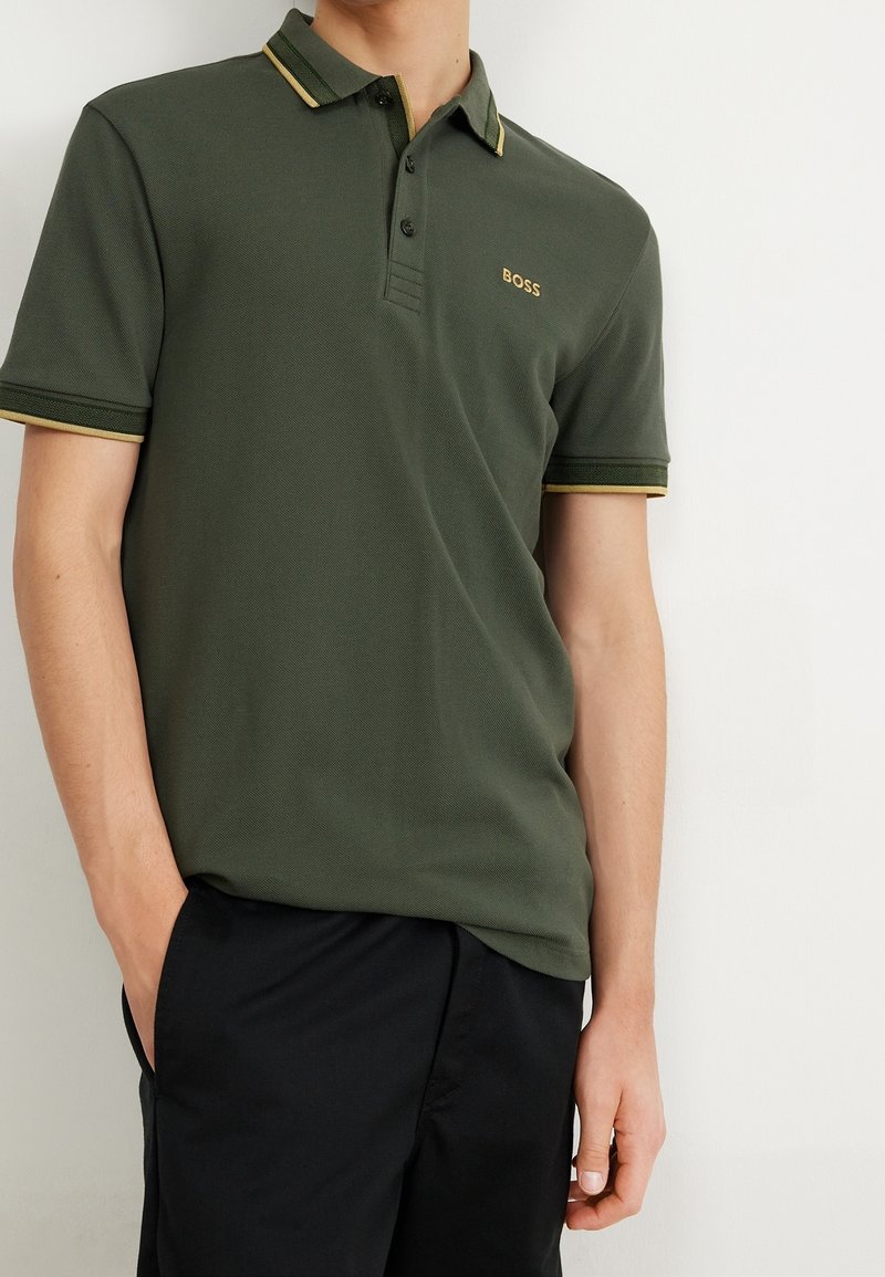 Mörkgrön polo skjorta med ribbad krage och gula detaljer, treknappsstängning och broderad logga. Bärs med svarta byxor.