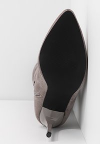 Zapato de tacón alto de ante gris con punta afilada, que presenta una suela negra brillante y lisa, y un pequeño grip de goma en el tacón.