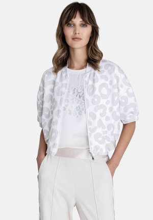 Vrouw met golvend haar die een witte cropped jas met een rits draagt met lichtgrijze luipaardvlekken, een wit grafisch T-shirt en witte broek, handen in de zakken.