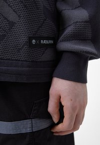 Personne portant un pull sombre à motifs avec une étiquette Timberland x RÆBURN, main posée près d’un pantalon noir avec une poche et une sangle visibles.