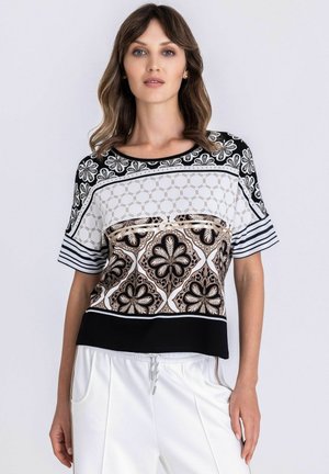 Femme portant un haut à manches courtes à motifs floraux et géométriques noirs, blancs et beiges, associé à un pantalon blanc à cordon de serrage.