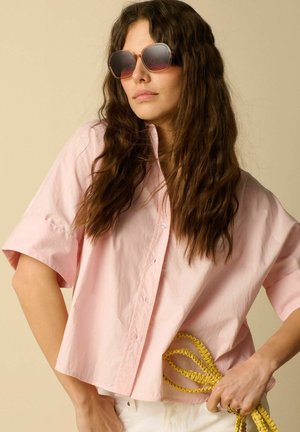 Vrouw met lang golvend haar die oversized zonnebrillen draagt en een los, lichtroze shirt met korte mouwen, die een geel gevlochten touw vasthoudt.