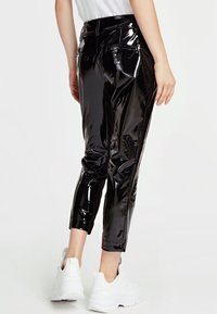 Glanzende zwarte cropped vinyl broek gedragen met witte zware sneakers, gezien van achteren op een persoon die tegen een witte achtergrond staat.