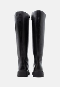 Ζευγάρι ψηλών μαύρων δερμάτινων μπότες με επίπεδες σόλες, φερμουάρ στη μέσα πλευρά, εμφανίζεται από το πίσω μέρος σε απλό φόντο.