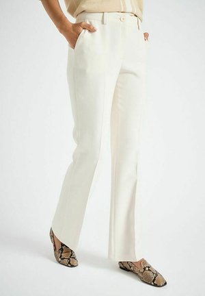 Persoon met een witte broek met rechte pijpen en hoge taille, handen in de zakken, en loafers met slangenprint op een witte achtergrond.