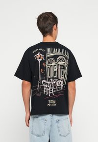 T-shirt nero a maniche corte con un design in linea di arte che rappresenta una scena di caffè con un lampione e posti all'aperto sul retro. Vestibilità casual.