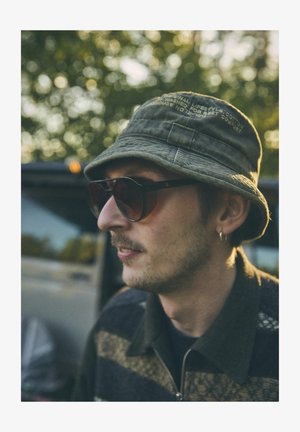 Klobúk typu bucket z olivovej bavlny, veľké slnečné okuliare s hnedým odtieňom a sveter so zipsom s hnedo-čiernymi horizontálnymi pruhmi.