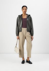 Giacca bomber in pelle verde oliva con cappuccio, abbinata a pantaloni beige a gamba larga e mocassini neri. Porta una borsa beige chiaro.