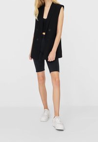 Black sleeveless blazer with a tailored fit, double buttons, worn over a black crop top and paired with black cycling shorts and white sneakers.