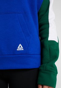 Blå huvtröja med framficka, gröna och vita ärmar samt en Reebok-logotyp på bröstet, tillverkad av mjuk bomullstyg.