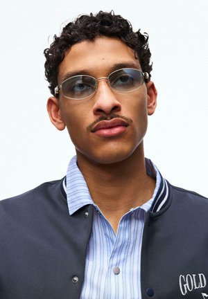 Mladi moški s kratkimi kodrastimi lasmi, ki nosi obarvana ovalna očala, modro srajco s črtami in temen suknjič z vezenim napisom "GOLD".