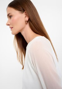 Witte sheer blouse met textuurstof, ronde halslijn en lange mouwen. Model draagt een gouden oorring. Bruin haar valt voorbij de schouders.