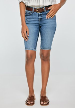 Kobieta ubrana w niebieskie krótkie spodenki dżinsowe do kolan, brązowy skórzany pasek, koszulę w paski oraz brązowe klapki na płaskim obcasie, stojąca na jednolitym tle.