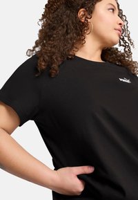 Sort bomulds t-shirt med rund hals og korte ærmer, med et lille hvidt logo på venstre brystområde. Glat tekstur.