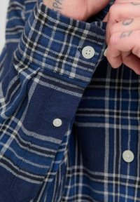 Detalle de una camisa de franela de cuadros en azul y blanco, que presenta un puño abotonado, una textura suave y acentos en negro en el patrón.