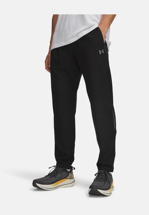 Mann trägt schwarze Under Armour Trainingshose und schwarze Laufschuhe mit gelben Akzenten, steht mit einer Hand in der Tasche.