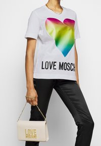 Bílé tričko s velkým barevným srdcem a textem "LOVE MOSCHINO". Kombinováno s černými kalhotami a béžovou kabelkou se zlatým nápisem.