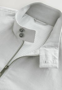 Chaqueta de cremallera gris claro con un collar estructurado, dos botones grises y un forro interior a rayas. Presenta una cremallera metálica y detalles cosidos.