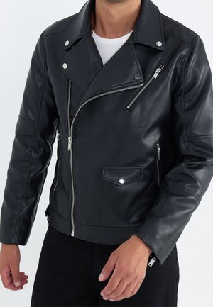 Mężczyzna w czarnej skórzanej kurtce motocyklowej z wieloma srebrnymi zamkami błyskawicznymi i zatrzaskami, w połączeniu z czarnymi spodniami i białą koszulą.