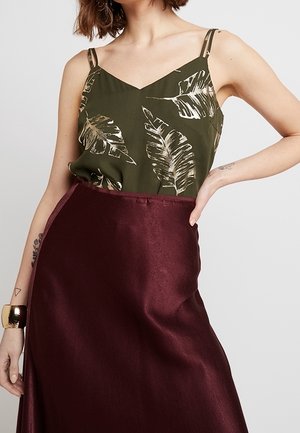 Žena nosí olivově zelený top s tenkými ramínky a béžovým listovým vzorem zastrčený do lesklé vínové sukně s vysokým pasem, ruka na boku.