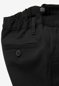 Un pantalon noir avec une taille élastique, une fermeture à bouton et une poche latérale avec un détail en bouton. Le tissu a une finition texturée.
