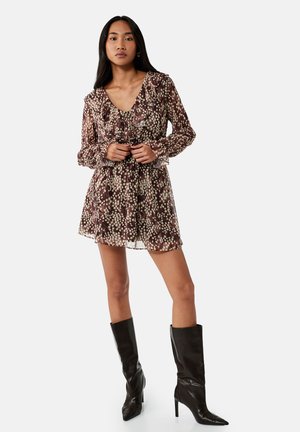 Vrouw staat met een bruin-wit gespikkelde, lange mouwen, met ruches mini-jurk en zwarte hooggehakte kniehoge laarzen op een witte achtergrond.