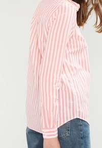 Ljusrosa och vita randiga skjortor med krage, långa ärmar och en rundad nederkant, kombinerad med blå jeans.