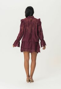Robe en dentelle bordeaux avec des manches à volants et une jupe superposée. Tissu transparent, col montant et détail en trou de serrure au dos.