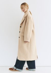 Beżowy, oversize płaszcz z teksturowanej tkaniny, z klapowanym kołnierzem, czarnymi guzikami i bocznymi kieszeniami, noszony z ciemnymi długimi spodniami i brązowymi butami.