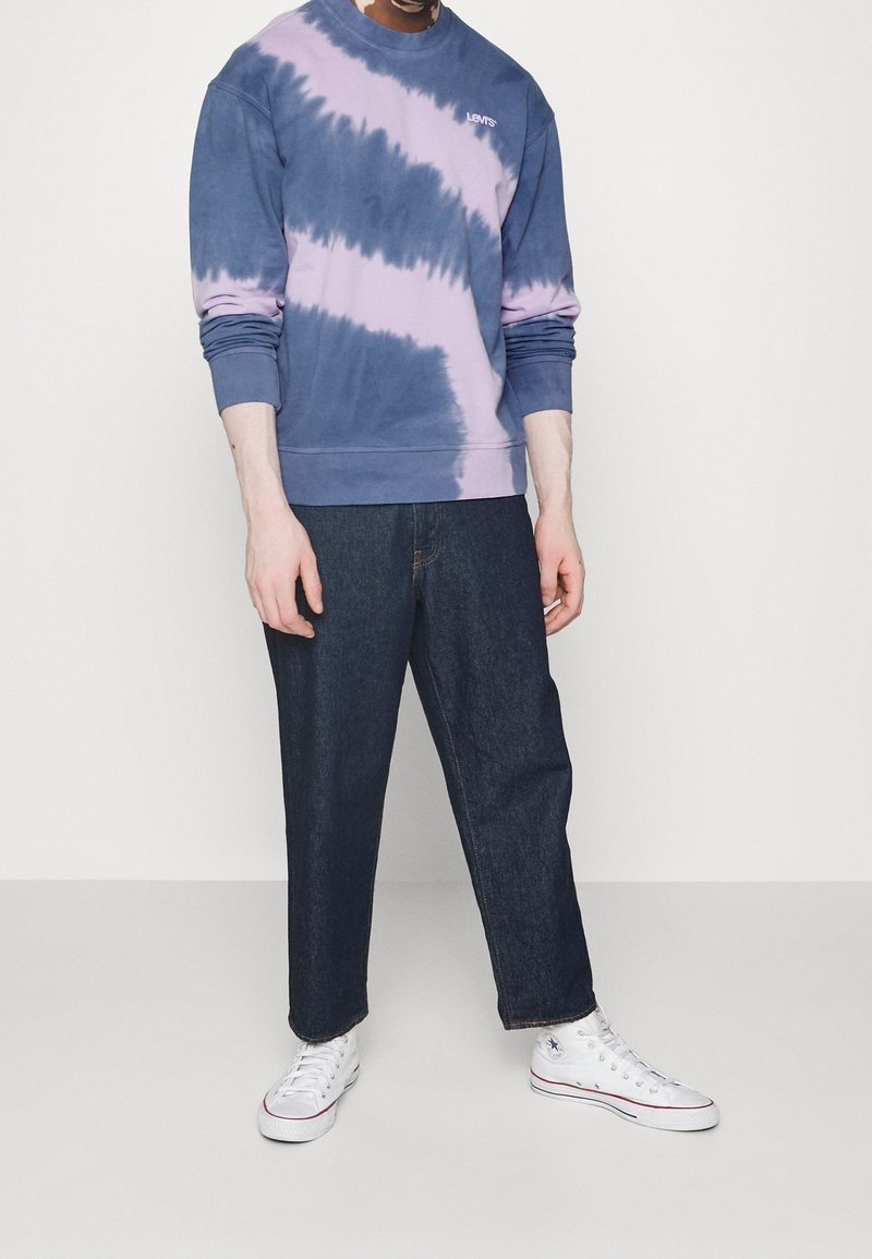 Felpa blu e viola con effetto tie-dye, colletto a girocollo, polsini a costine e logo in rilievo; abbinata a pantaloni in denim scuro e sneakers bianche.