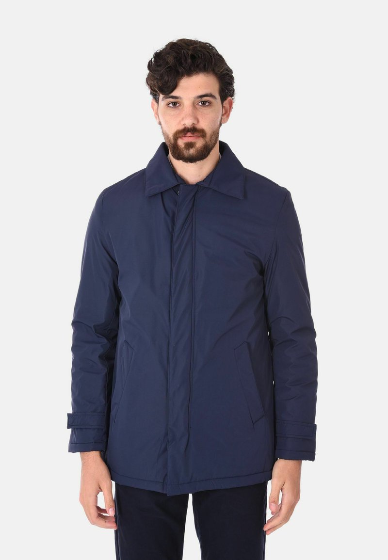 Cappotto isolato blu navy con colletto, chiusura frontale con bottoni, tasche laterali e texture imbottita, indossato da un uomo con capelli e barba scuri.