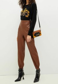 Maglione nero con design grafico oro, pantaloni a vita alta in finta pelle marrone e stivaletti neri con punta affilata e catene.