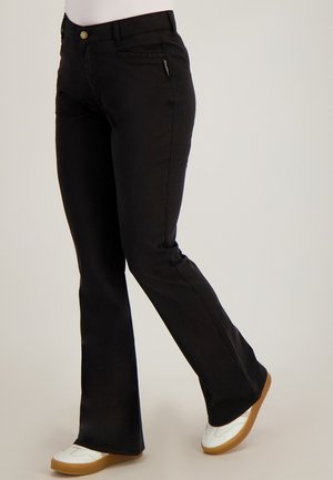 Persoon draagt zwarte bootcutbroek en witte sneakers met rubberen zolen, staand tegen een effen lichte achtergrond.