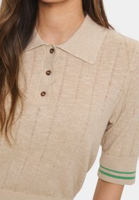 Beige short-sleeve polo shirt with a textured pattern, scalloped collar, and three brown buttons. Ribbed cuff features green stripes.