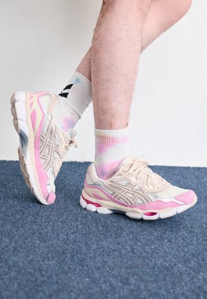 Legs wearing white and pink running shoes and white tie-dye socks standing on blue carpet against a white wall.