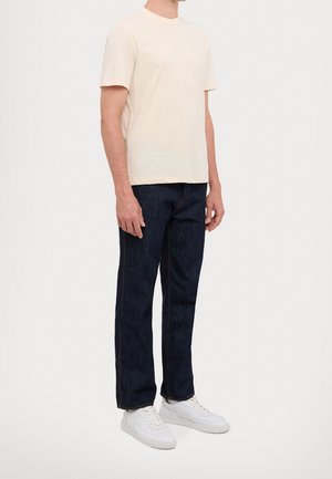 Homme portant un t-shirt uni beige clair à manches courtes, un jean bleu foncé et des baskets blanches, debout devant un fond blanc.