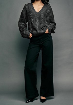 Femme portant un pull en maille torsadée gris foncé à col en V, un pantalon noir taille haute à jambes larges et des talons pointus noirs, les mains dans les poches.