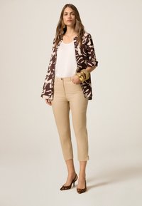 Braun und beige gemusterte Jacke über einem weißen Oberteil, kombiniert mit beigen, knöchellangen Hosen und Leopardenmuster-Absätzen, versehen mit goldenen Accessoires.